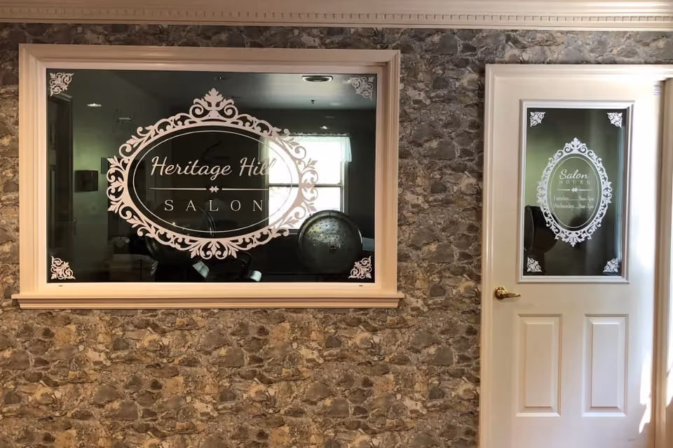 Interior view of a salon at Heritage Hill Senior Community showing a stone wall with a large window and a white door. Both the window and door have decorative white decals with the salon's name and hours of operation.