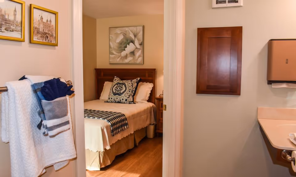 View through a doorway showing a bedroom with a neatly made bed featuring decorative pillows and a blanket. The bedroom has a wooden headboard and a floral painting above it. In the foreground, there is a bathroom area with a towel rack holding folded towels and washcloths, a wooden cabinet mounted on the wall, and a paper towel dispenser next to a sink.