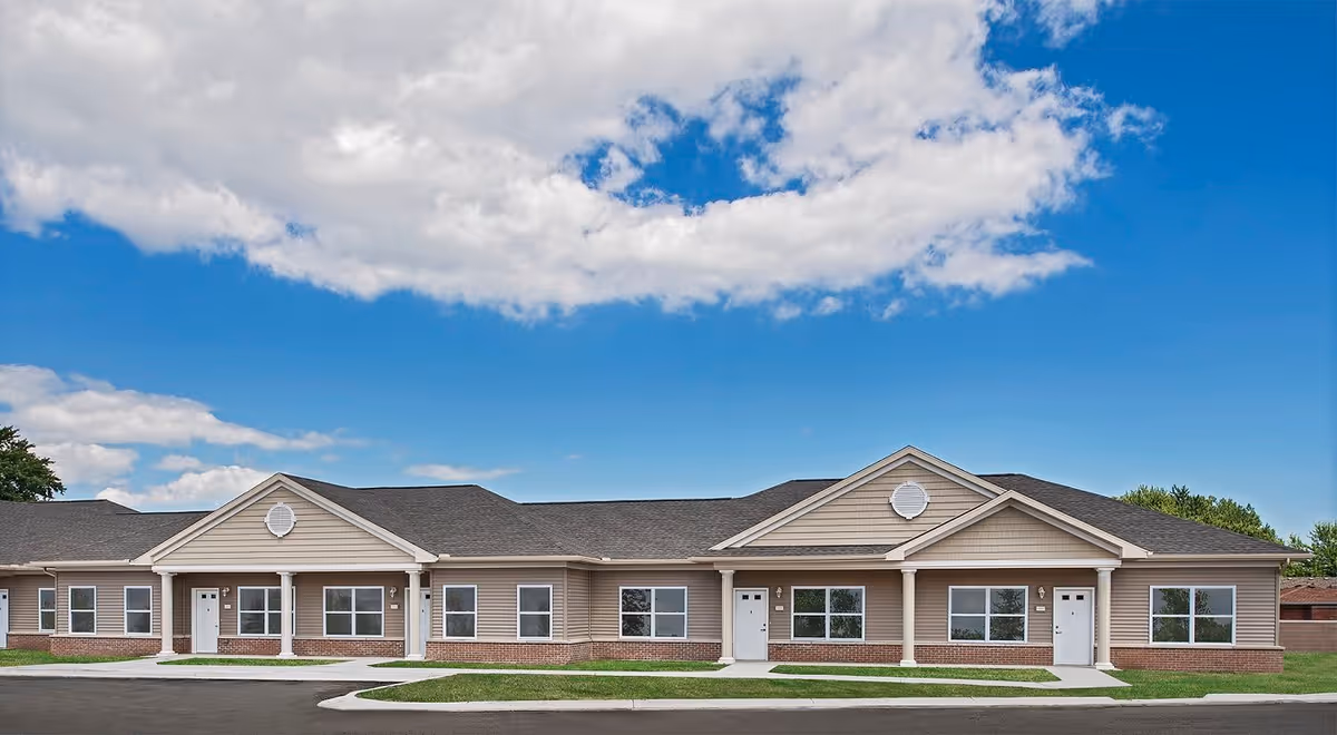 Exterior view of a single-story residential building with beige siding, brick accents, and multiple white doors and windows under a partly cloudy blue sky.