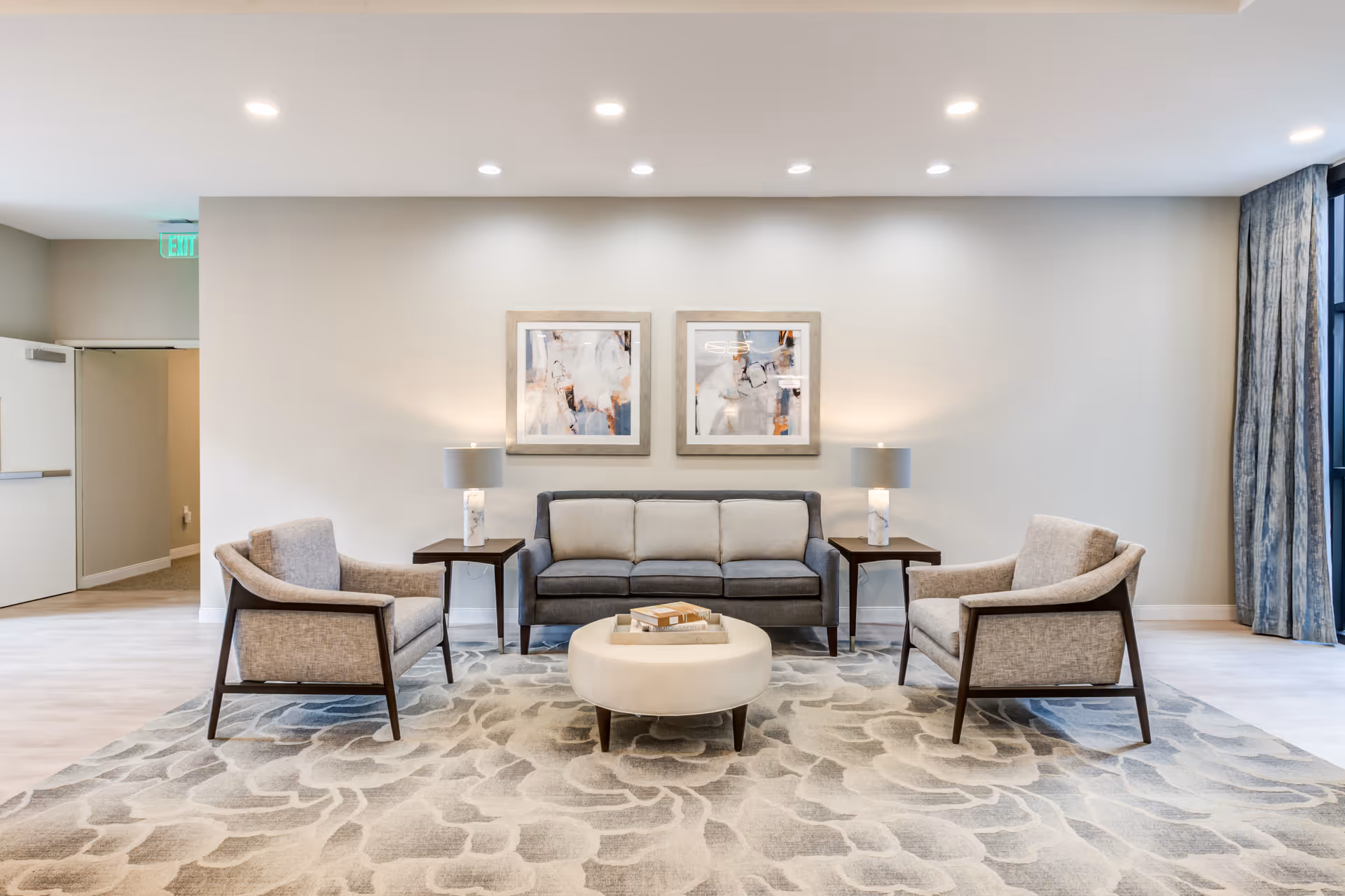 A modern living room area with a gray sofa, two beige armchairs, a round beige ottoman, two side tables with lamps, abstract artwork on the wall, and a patterned area rug on a light wood floor.