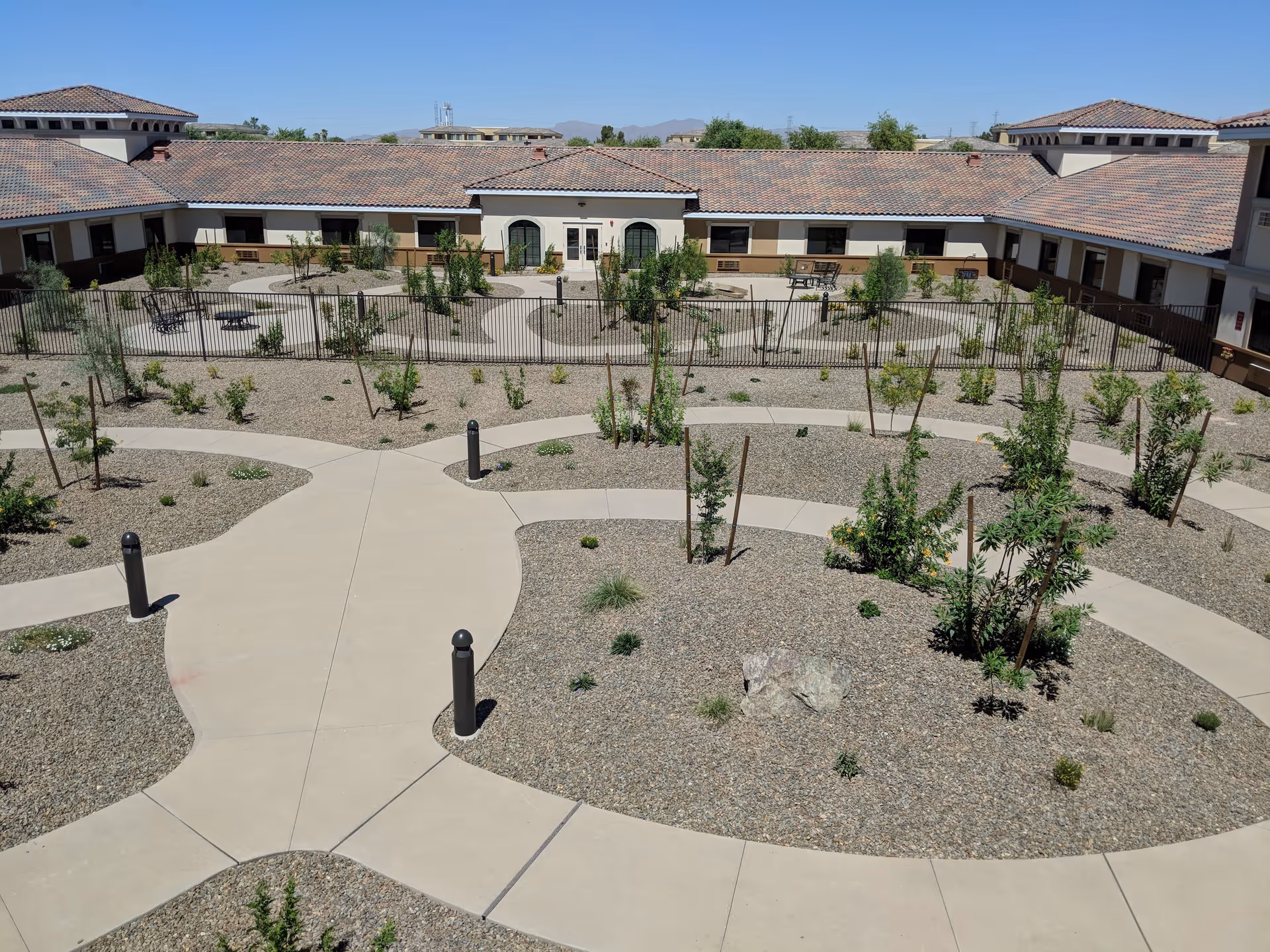 Outdoor courtyard area of The Mission at Agua Fria Senior Living featuring paved walkways, small trees and shrubs planted in gravel beds, benches, and a fenced section with additional seating. The building surrounds the courtyard with a tiled roof and multiple windows under a clear blue sky.