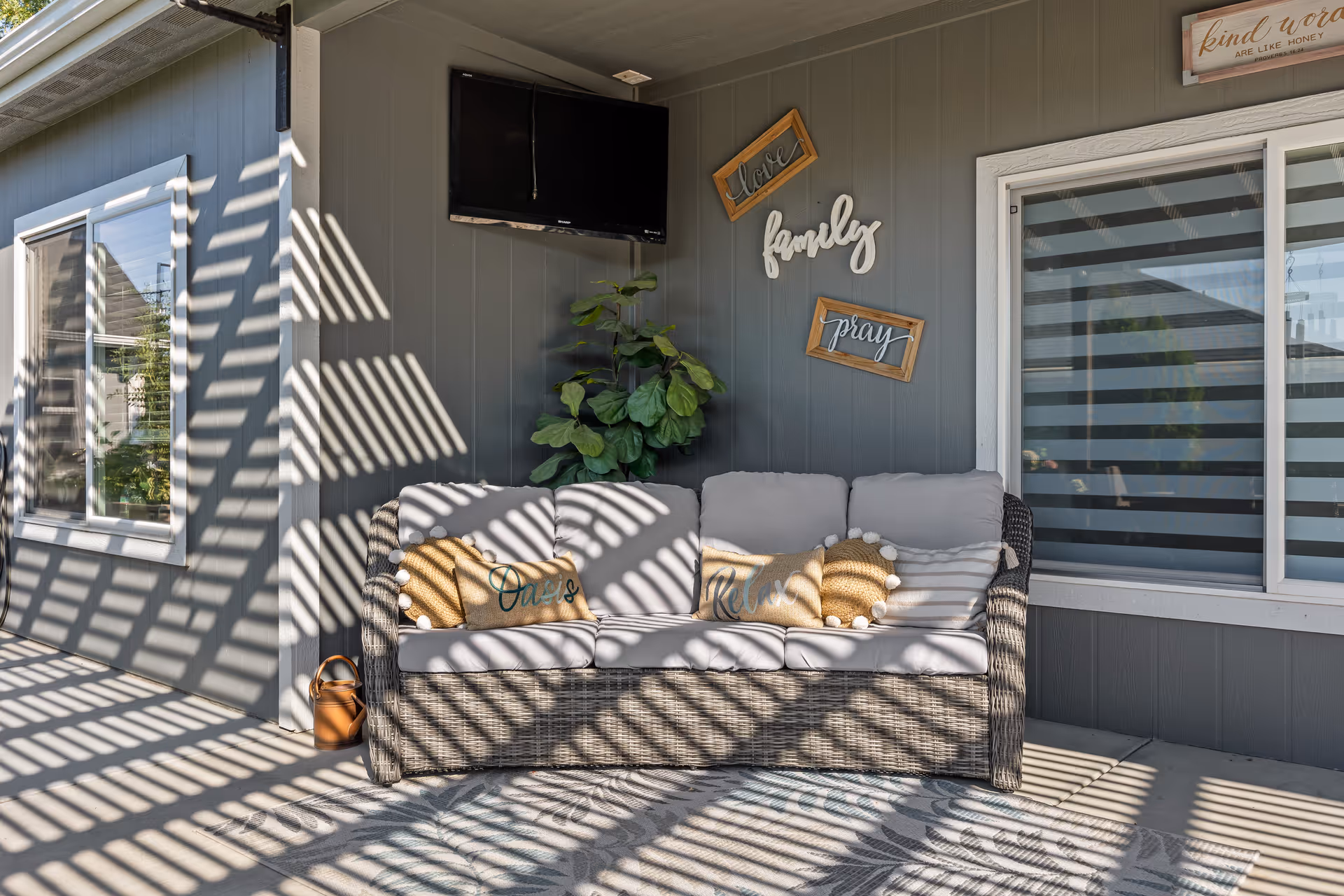 Sunlit covered patio with a wicker sofa, decorative pillows, wall decor reading 'family' and 'pray', a potted plant and a mounted TV.