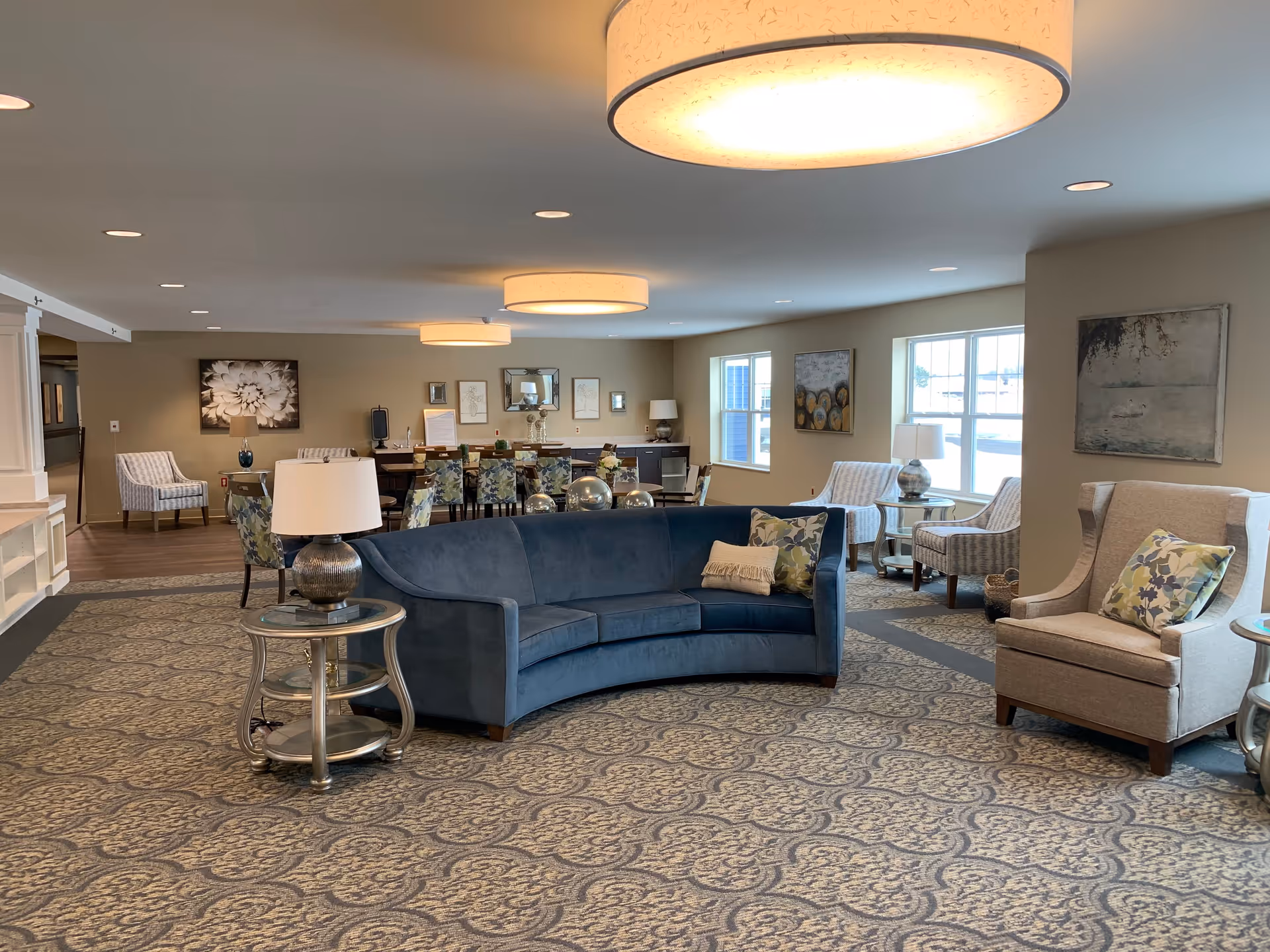 A spacious senior living community common area featuring a curved blue sofa with decorative pillows, beige armchairs, side tables with lamps, patterned carpet, and a dining area with multiple chairs and tables in the background. The room is well-lit with large windows and ceiling lights, and decorated with framed artwork on the walls.