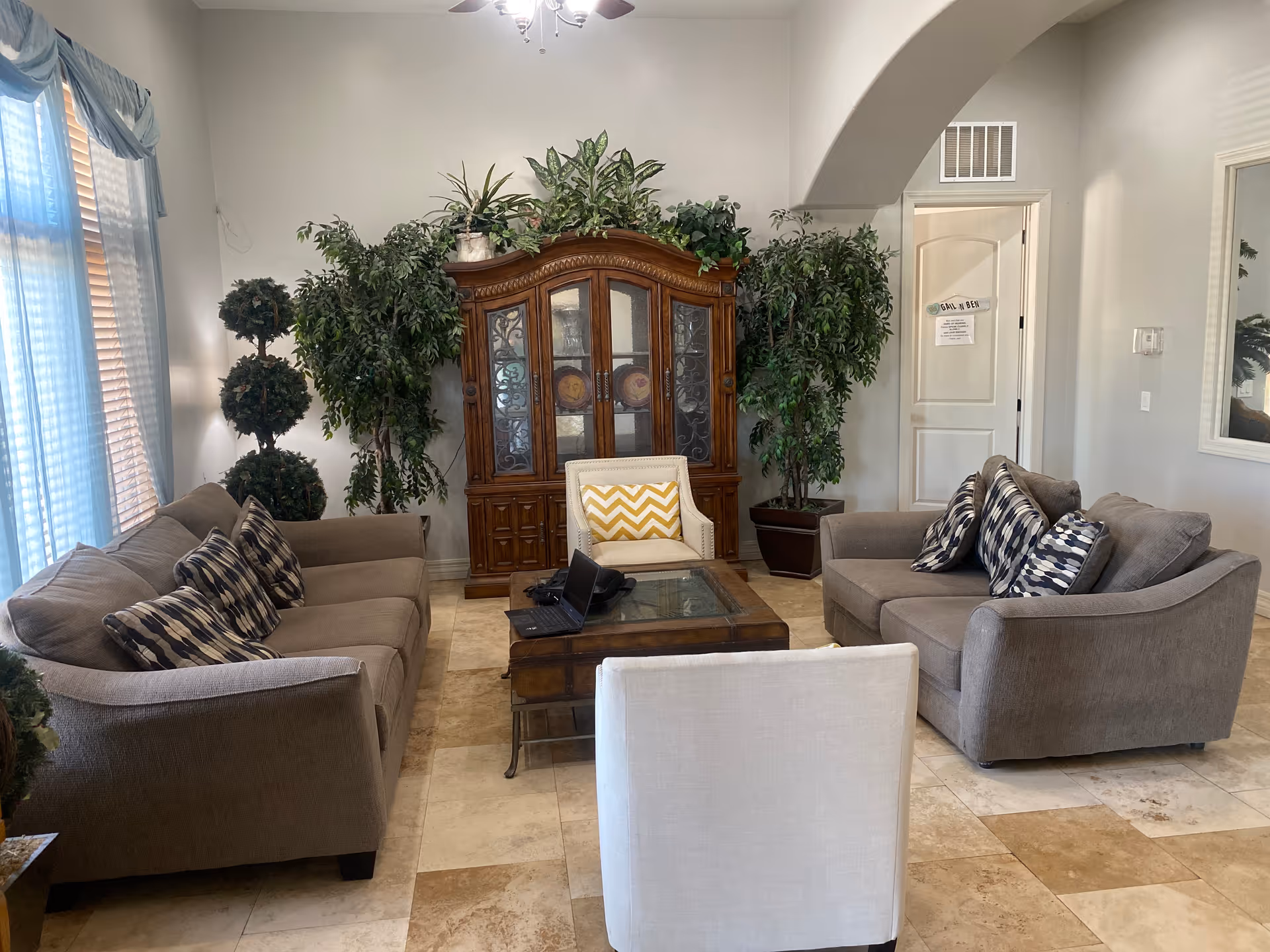 Furnished common sitting room with two gray sofas, chairs, a wooden display cabinet, coffee table, and potted plants.