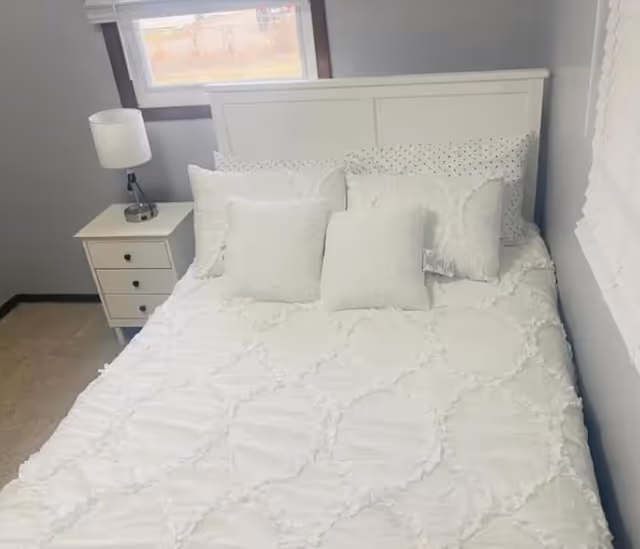 A neatly made bed with a white headboard, white textured bedspread, and multiple white pillows. To the left of the bed is a small white nightstand with three drawers and a white lamp on top. There is a window above the nightstand and another window with white blinds on the right wall.