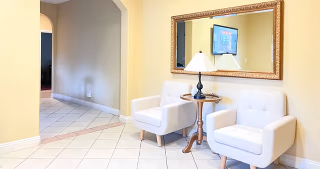 A bright interior space with two white cushioned armchairs positioned around a small wooden side table with a lamp. A large ornate mirror hangs on the yellow wall behind the chairs, reflecting a mounted flat-screen TV. The floor is tiled with white and beige tiles, and an arched doorway leads to another room.