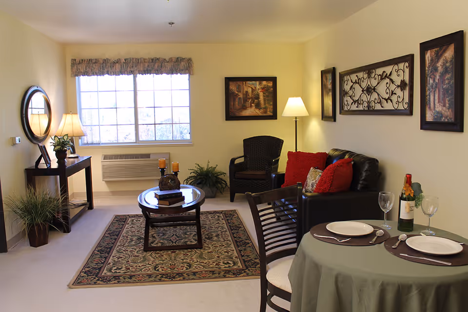 A cozy living room with a large window letting in natural light. The room features a dark leather couch with red and patterned pillows, a wicker chair, a round wooden coffee table with candles and books, and a patterned area rug. There is a small dining table set for two with plates, utensils, wine glasses, and a bottle of wine. The walls are decorated with framed artwork and a decorative metal wall hanging. A floor lamp and a table lamp on a console table provide additional lighting. Several plants add greenery to the space.