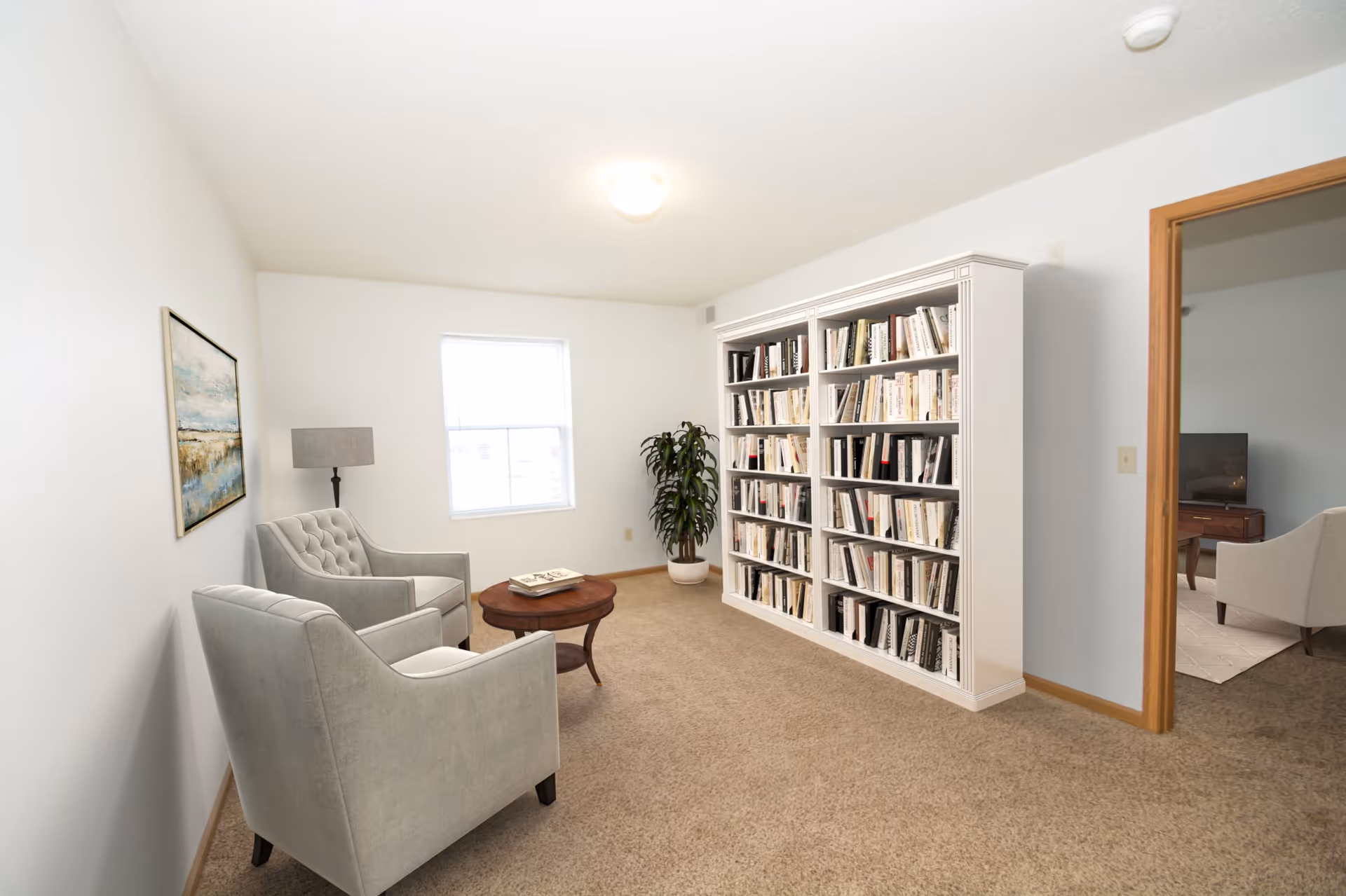 A cozy reading room with two gray upholstered armchairs, a round wooden coffee table with a book on it, a large white bookshelf filled with books, a potted plant, a window letting in natural light, and a framed landscape painting on the wall. There is an open doorway leading to another room with a TV and additional seating.