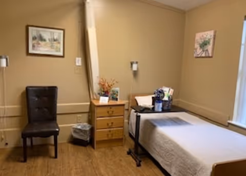 A simple nursing facility room with a single bed covered with a white blanket, a wooden nightstand with drawers, a black chair, a small trash bin, and two framed pictures on beige walls. There is a window letting in natural light on the right side.
