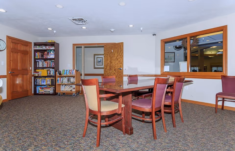 A room with a rectangular wooden table surrounded by six chairs, some with purple upholstery and one with beige upholstery. The room has carpeted flooring, a wooden door, a bookshelf filled with books and games, and a window looking into another room.