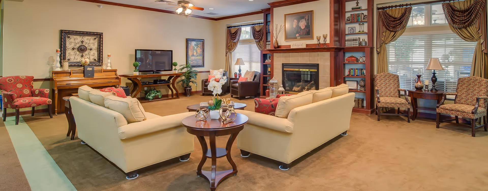 A cozy living room with two beige sofas arranged around a wooden coffee table with decorative items. The room features a fireplace with built-in bookshelves on either side, a piano with candles and decorative pieces, a TV on a wooden stand, and several armchairs near windows with draped curtains. The walls are painted light beige, and the carpet is brown.