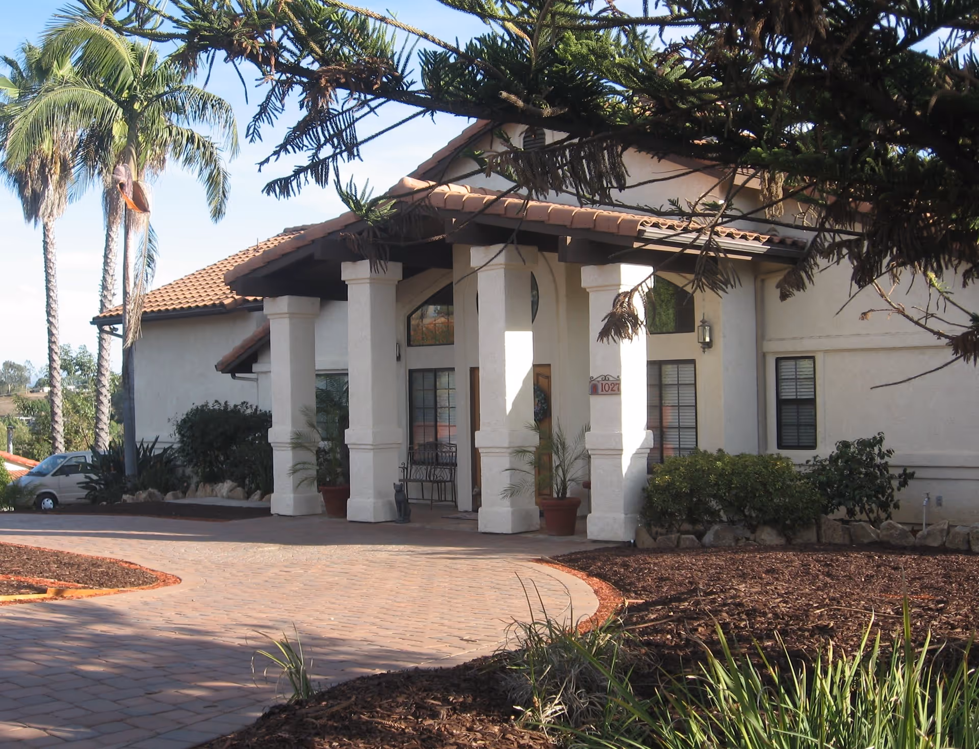 Exterior view of a single-story building with a tiled roof and white columns at the entrance. There are palm trees and landscaped plants around the paved driveway leading to the entrance.