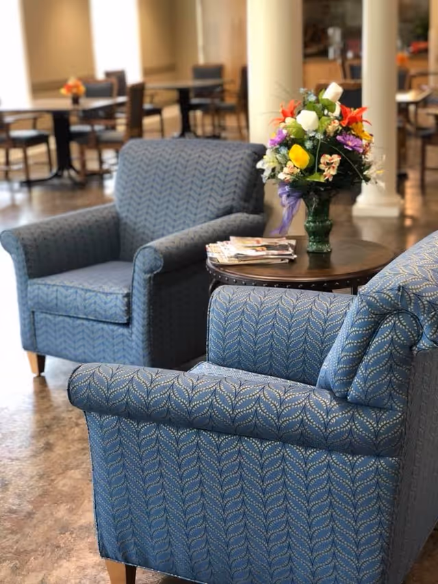 Two blue patterned armchairs positioned around a small round wooden table with a colorful floral arrangement and a stack of magazines, set in a spacious room with additional tables and chairs in the background.