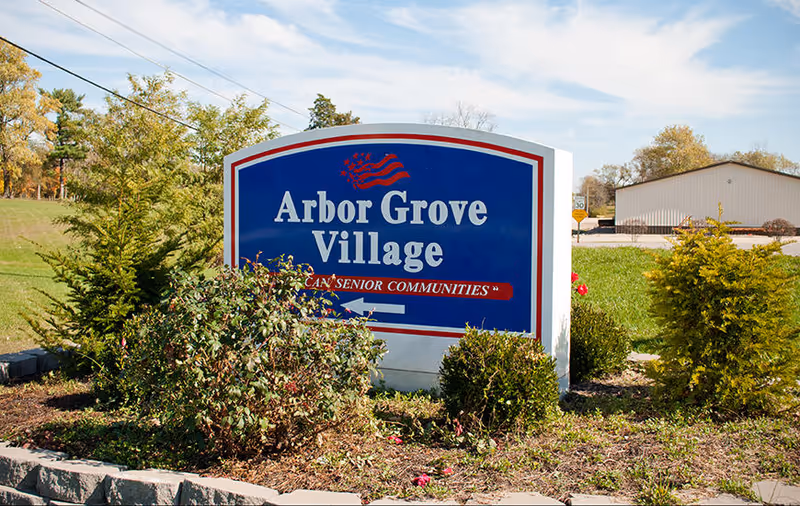 Outdoor view of a blue and white sign for Arbor Grove Village, surrounded by bushes and small trees, with a building and grassy area in the background under a partly cloudy sky.
