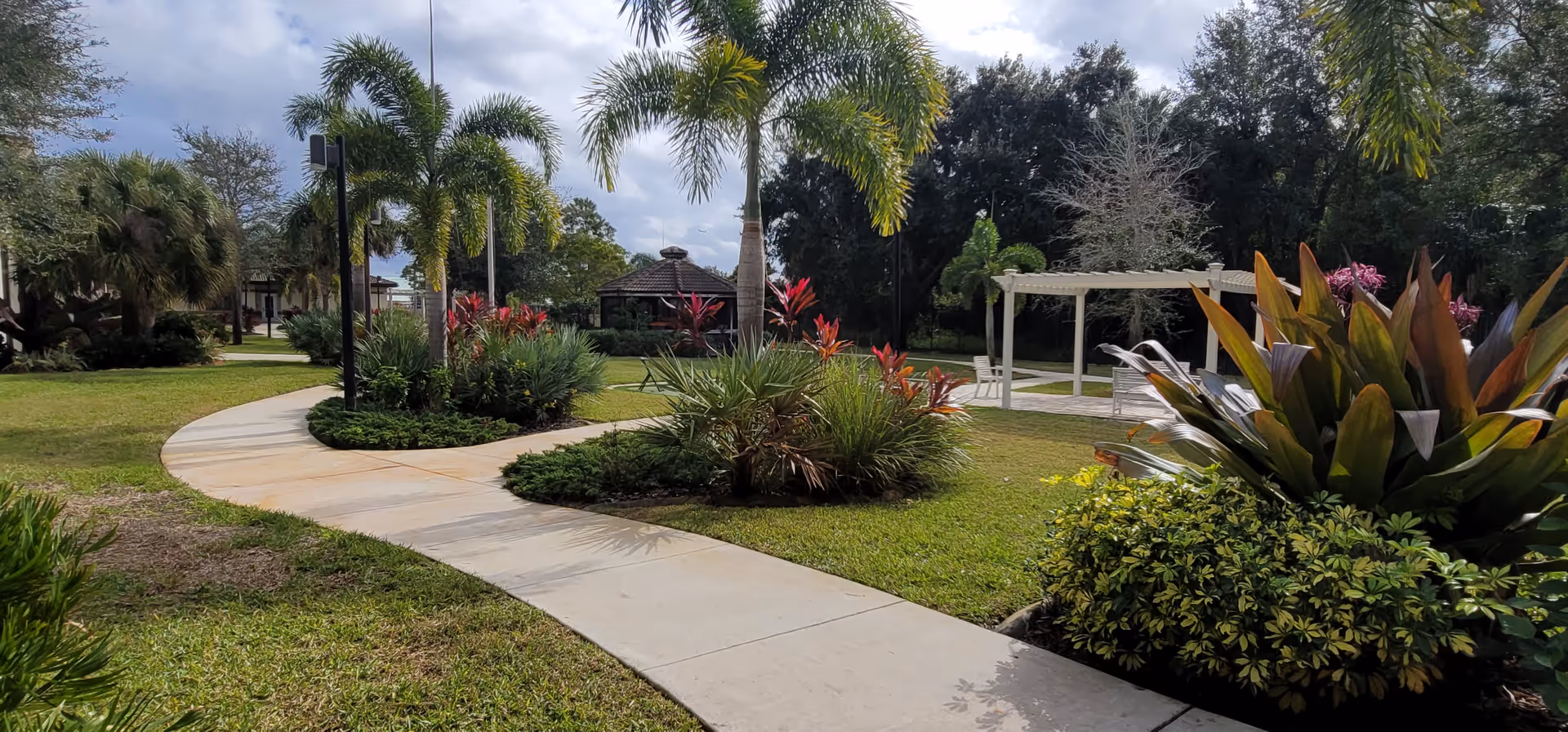 A landscaped outdoor garden area with a curved concrete walkway, palm trees, various shrubs, and flowering plants. There is a gazebo in the background and a white pergola with seating on the right side, under a partly cloudy sky.