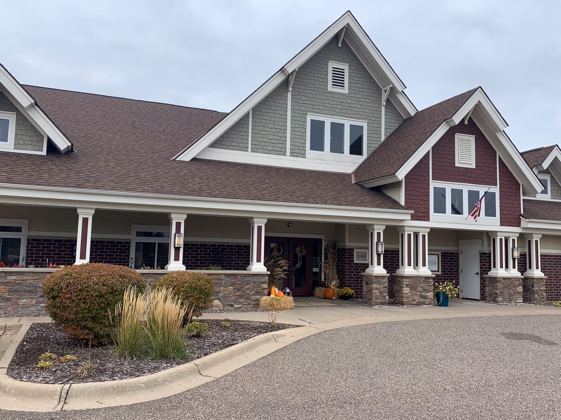 Front exterior view of a senior living facility building with a covered entrance, stone and brick facade, and decorative plants and pumpkins near the doorway under a cloudy sky.