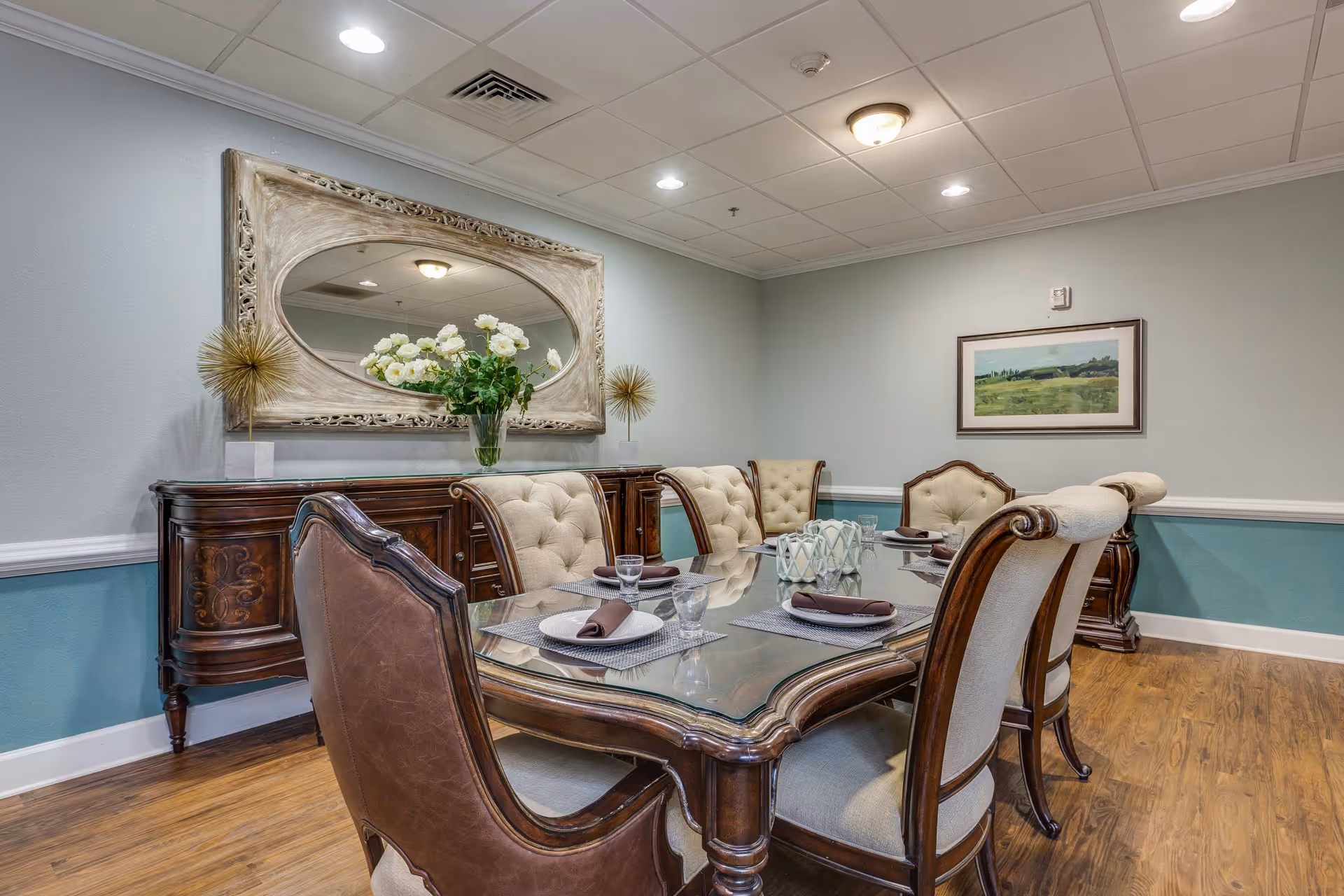 A formal dining room with a wooden table topped with glass and six upholstered chairs. The table is set with plates, glasses, and brown napkins. A wooden sideboard against the wall holds a vase with white flowers and decorative golden spiked ornaments. Above the sideboard is a large ornate mirror. The walls are painted light gray with a teal wainscoting, and a framed landscape painting hangs on the adjacent wall. The floor is wood, and the ceiling has recessed lighting and a central light fixture.