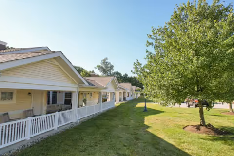 Row of single-story cottages with covered porches facing a green lawn and trees under a clear blue sky.