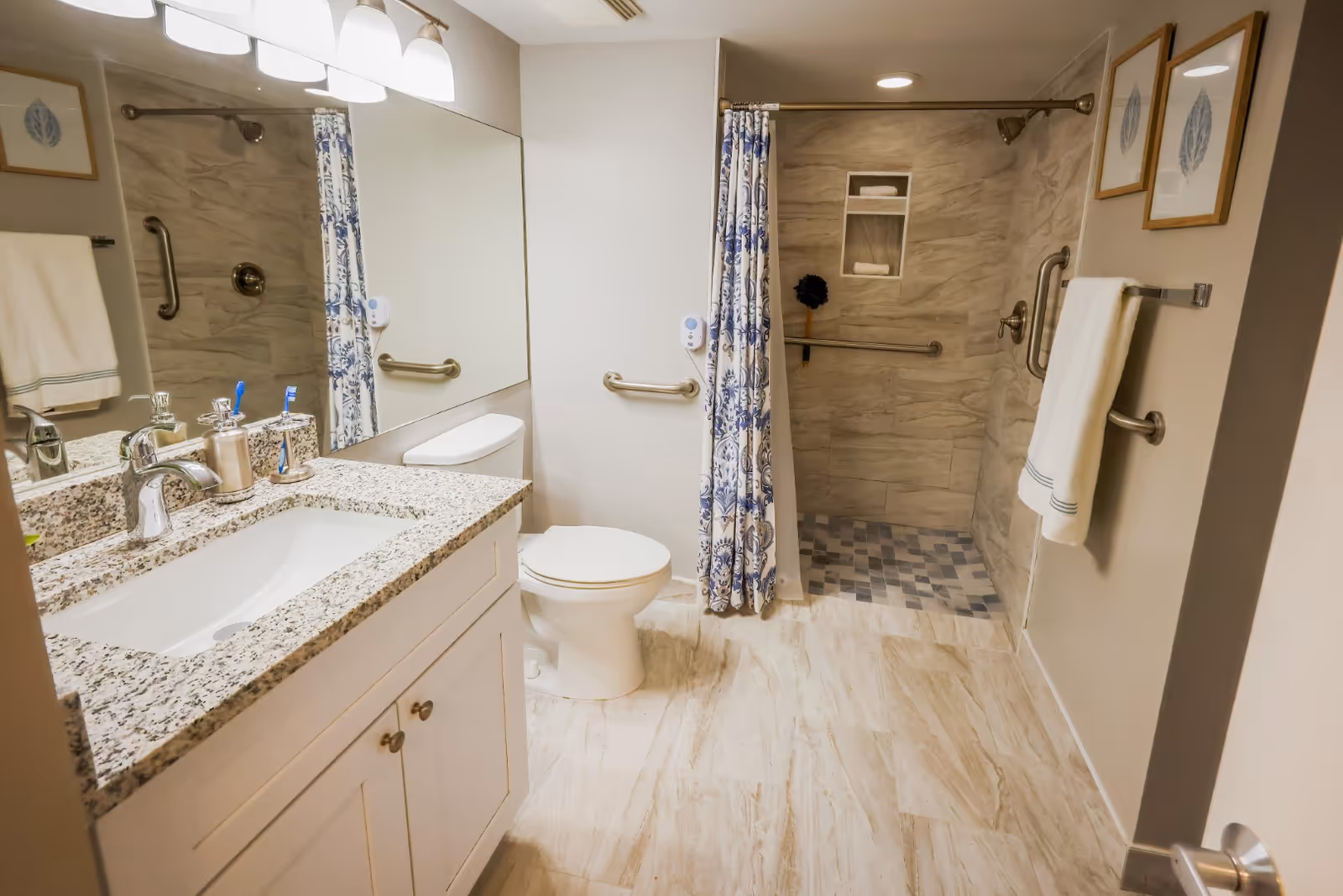 Accessible bathroom with a granite-topped vanity and sink, toilet, and a roll-in tiled shower with grab bars and a patterned curtain.