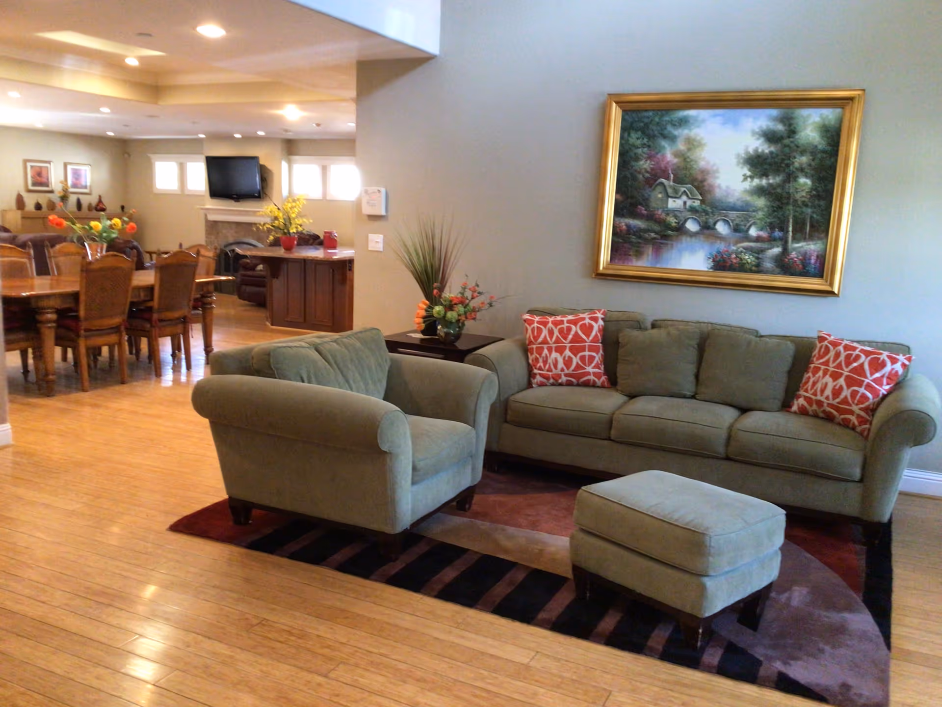 A cozy living room area with a green sofa, matching armchair, and ottoman on a patterned rug. The sofa has two red and white patterned throw pillows. A framed painting of a scenic landscape with a house, bridge, and trees hangs on the wall above the sofa. In the background, there is a dining area with a wooden dining table and chairs, a fireplace with a TV mounted above it, and decorative flowers on tables.