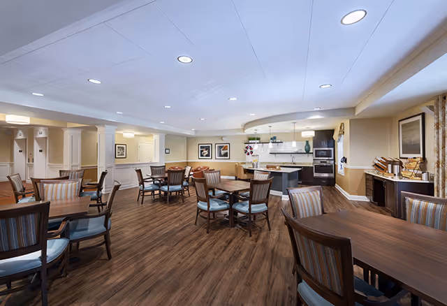 A spacious dining area with multiple wooden tables and chairs arranged neatly on a wood floor. The room has a modern kitchen area in the background with an island, oven, and cabinets. The walls are painted beige with white trim, and there are framed pictures and light fixtures on the walls and ceiling.
