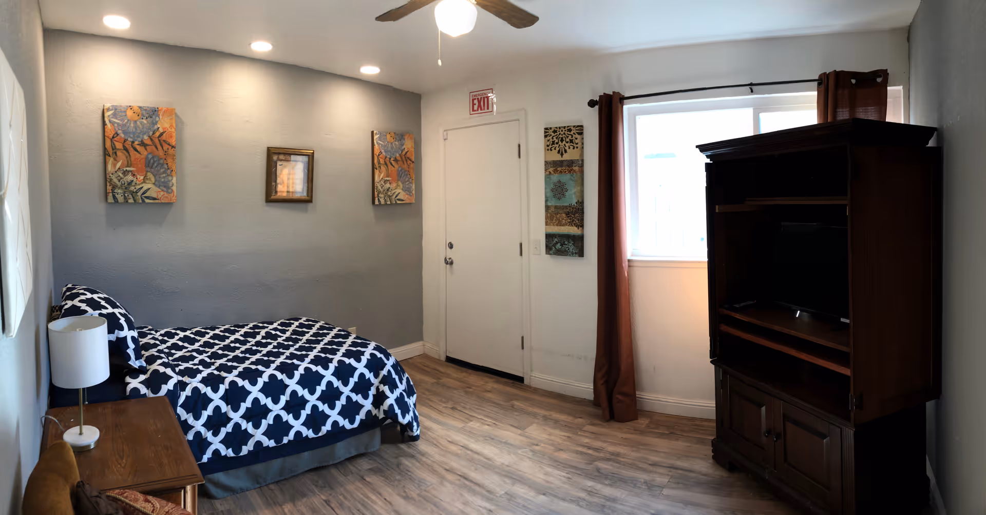 A small bedroom with a single bed covered in a blue and white patterned bedspread. There is a wooden nightstand with a white lamp next to the bed. The walls are painted light gray and decorated with three framed artworks. A ceiling fan with a light is mounted on the ceiling. A window with brown curtains is on the right side, next to a wooden entertainment cabinet with a TV inside. The floor is wood laminate, and there is a white door with an exit sign above it.