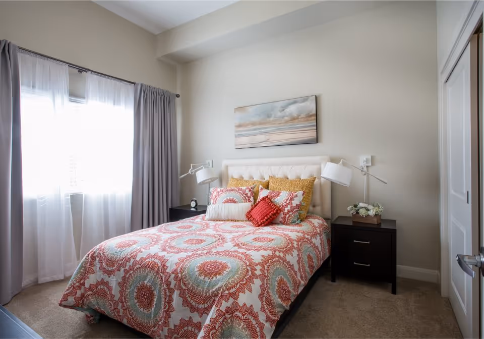 A cozy bedroom with a patterned bedspread, upholstered headboard, nightstands and lamps, a curtained window, and wall art above the bed.