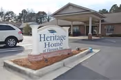 Exterior view of Heritage Home of Florence Inc, showing the facility's entrance with a covered drop-off area and a sign with the facility's name in front. A white vehicle is parked nearby and trees are visible in the background.