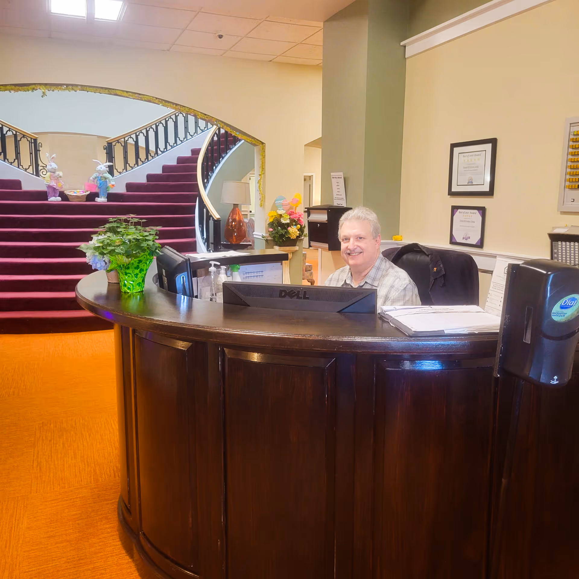 Reception area with a smiling man sitting behind a curved wooden desk. Behind him is a staircase with red carpet and decorative bunny figures on the steps. The area is well-lit with a plant on the desk and framed certificates on the wall.