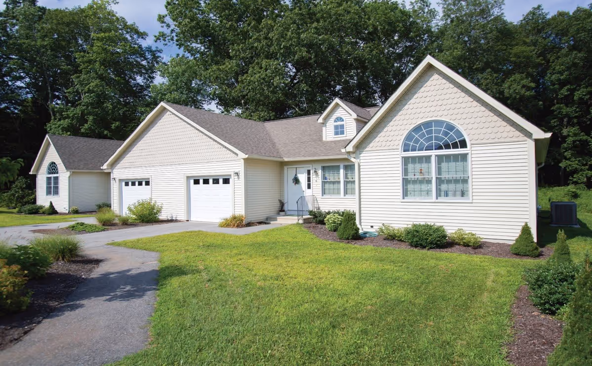 Exterior view of a single-story residential building with beige siding, a two-car garage, a front door with a small set of stairs, and large windows. The building is surrounded by a well-maintained lawn, shrubs, and trees in the background under a clear sky.