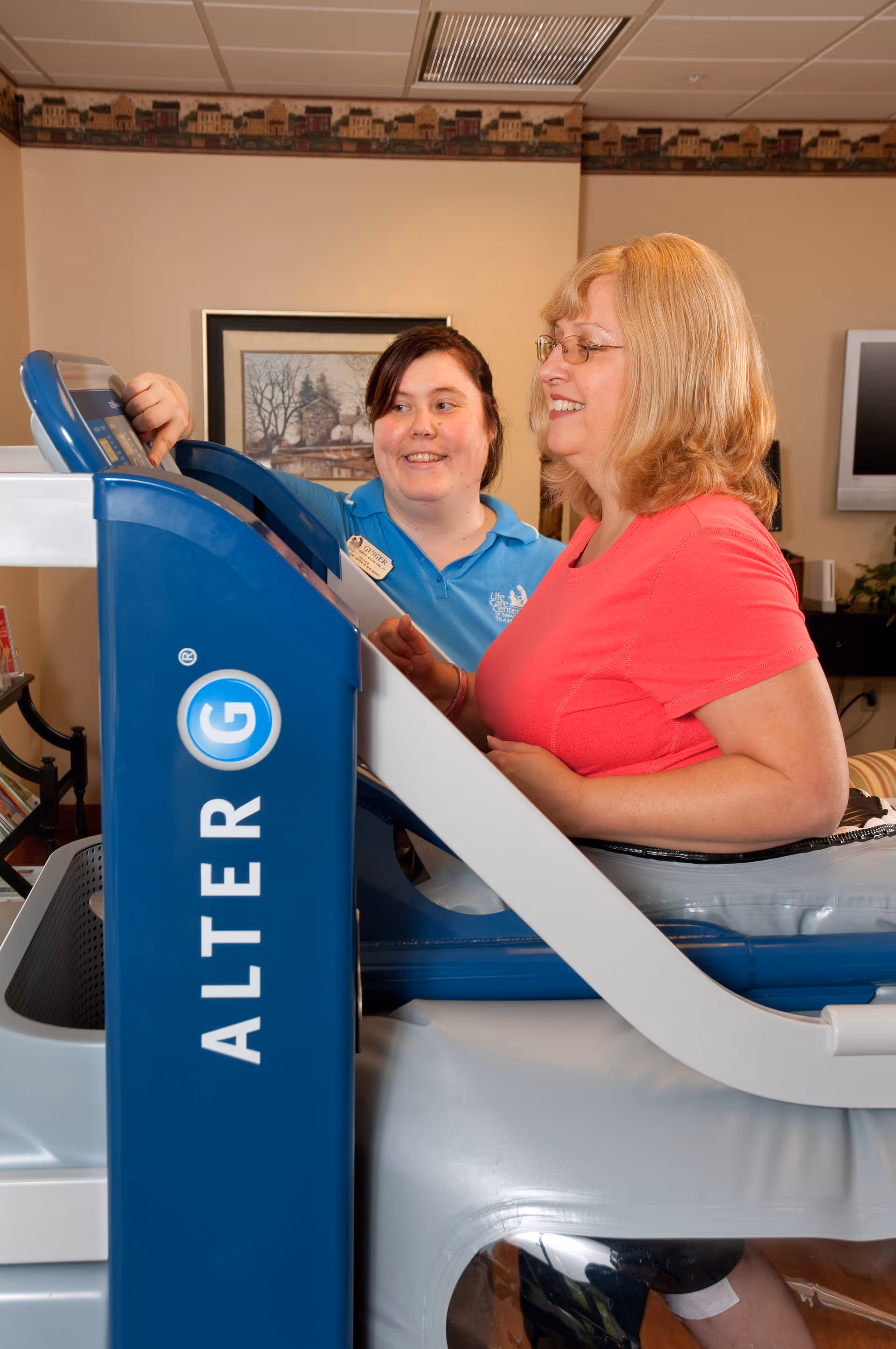 A woman in a pink shirt is using an AlterG anti-gravity treadmill while a staff member in a blue shirt assists her. They are indoors in a room with beige walls, a framed picture, and a television in the background.