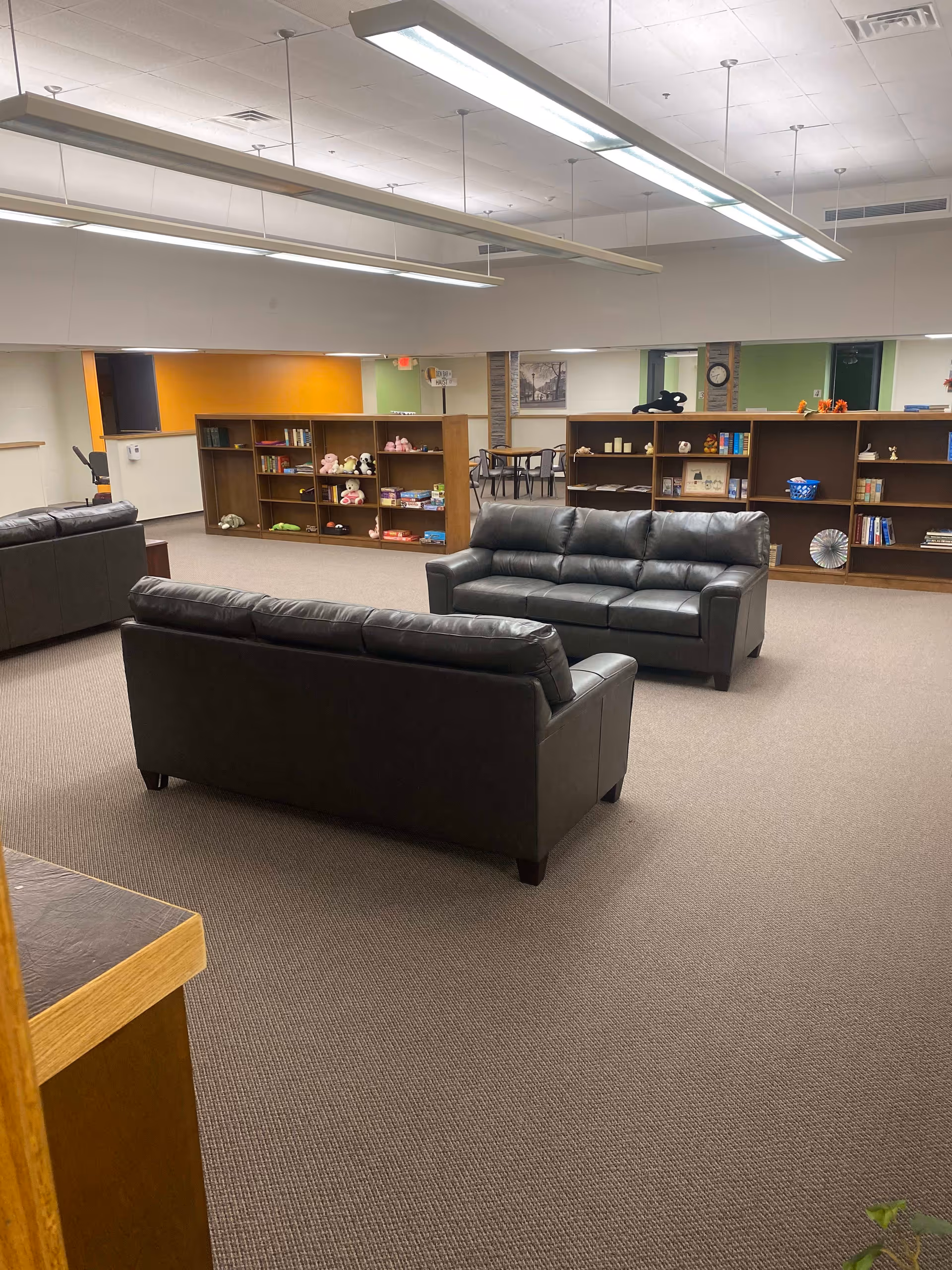 A spacious indoor common area with two black leather sofas facing each other on a carpeted floor. Behind the sofas are wooden bookshelves filled with books, toys, and decorative items. In the background, there is a small table with chairs and a wall with a clock and framed pictures. The ceiling has long fluorescent light fixtures.