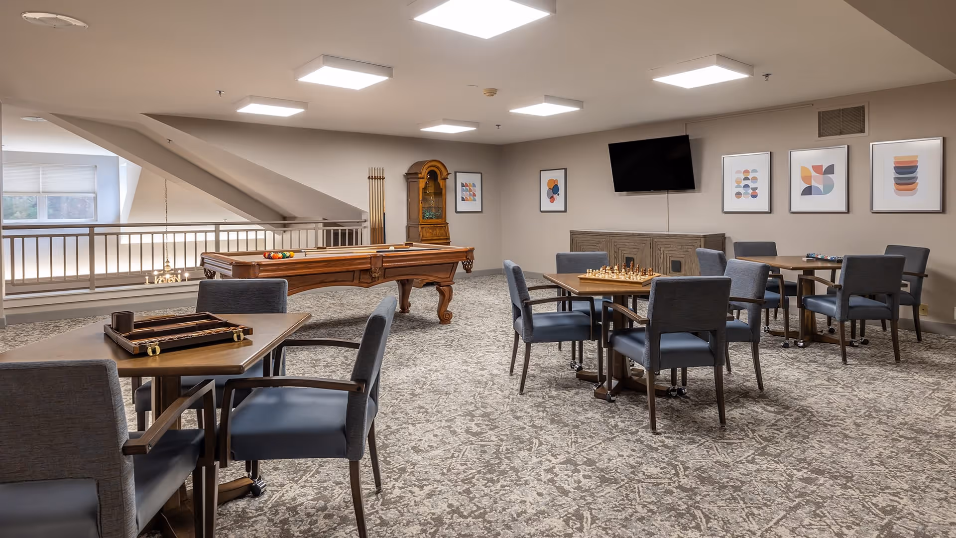 A spacious game room with several tables and chairs arranged for playing board games like chess and backgammon. There is a pool table with billiard balls set up, a grandfather clock, and a wall-mounted TV. The room has carpeted floors, neutral-colored walls, and framed abstract artwork on the walls.