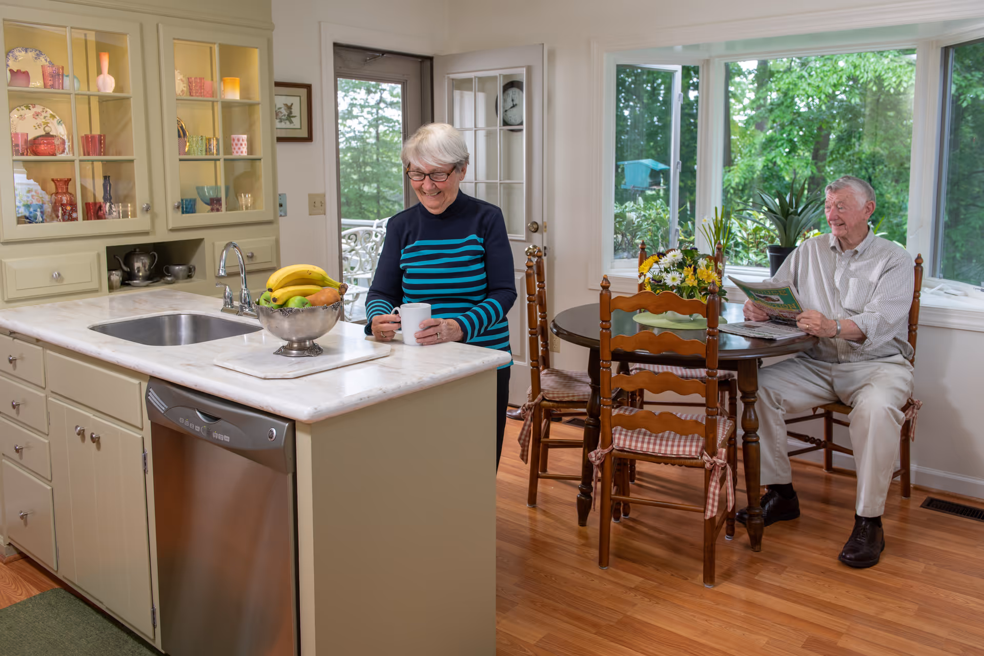 An elderly woman standing in a kitchen holding a white mug, smiling. An elderly man is sitting at a round wooden dining table with a floral centerpiece, reading a newspaper. The kitchen has light-colored cabinets, a stainless steel dishwasher, a sink, and a bowl of fruit on the counter. Large windows behind the man show green trees outside.