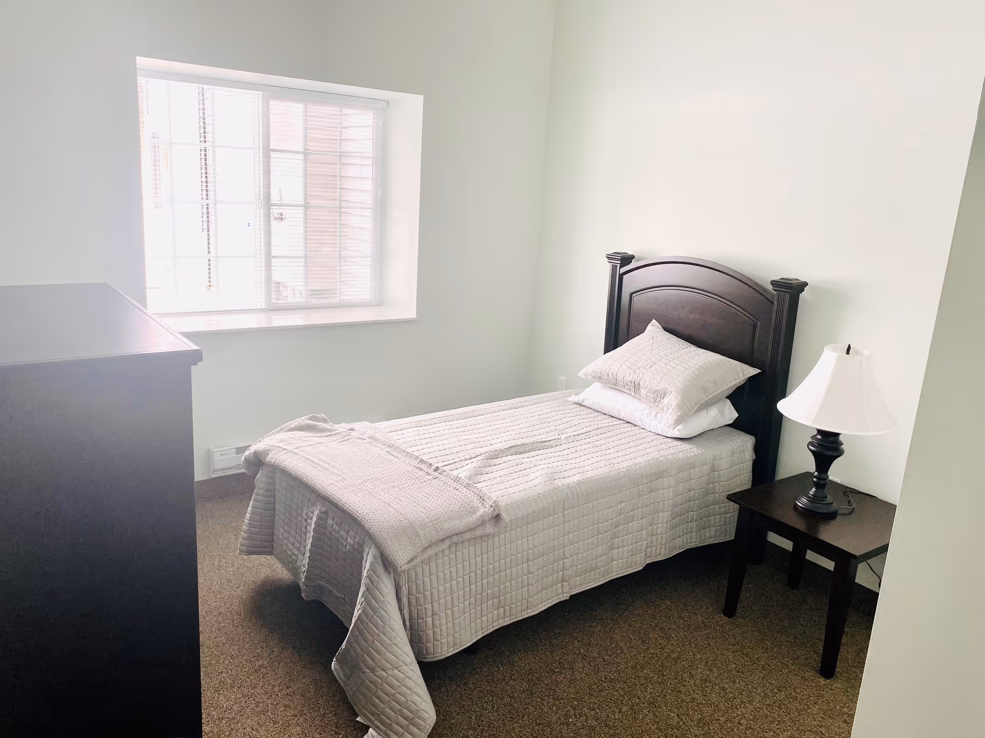 A small, simple bedroom with a single bed covered in a light gray quilt and two pillows. Next to the bed is a dark wooden nightstand with a white lampshade. There is a window with blinds letting in natural light, and a dark wooden dresser is partially visible on the left side of the image. The walls are white and the floor is carpeted in a neutral tone.