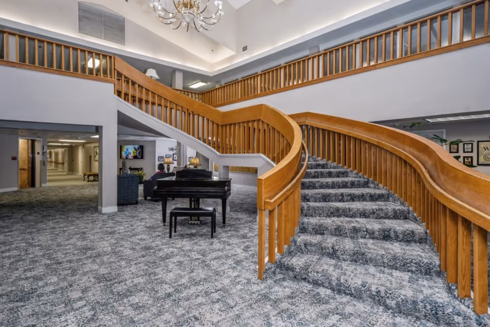 Interior view of Ashwood Place featuring a carpeted curved staircase with wooden railings, a black grand piano with a bench, a seating area with a person watching a wall-mounted TV, and a chandelier hanging from the ceiling.