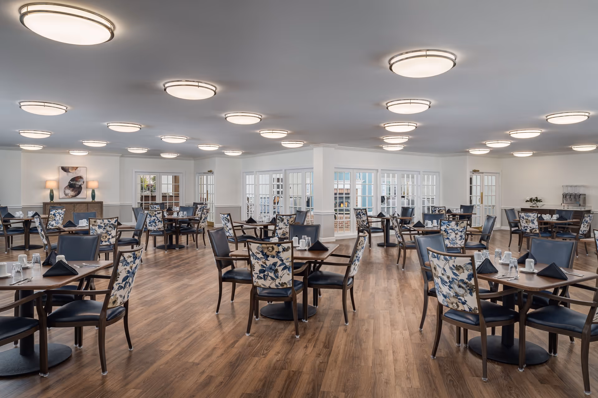 A spacious dining room with multiple wooden tables and chairs arranged neatly. The chairs have dark blue seats and floral-patterned backs. The room has wooden flooring, white walls, and numerous round ceiling lights. There are glass-paneled doors and windows along the walls, and a sideboard with lamps and artwork is visible in the background.