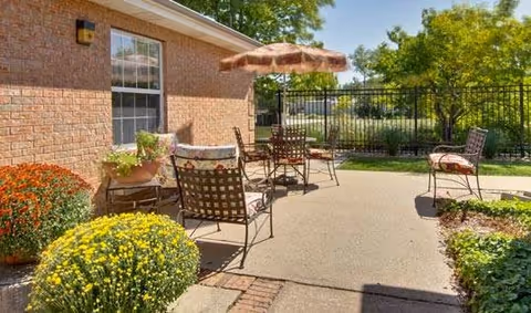 Outdoor patio area with metal chairs and tables, one table shaded by a large umbrella. The patio is adjacent to a brick building with windows. Surrounding the patio are various plants and flowers, including yellow and orange chrysanthemums. A black metal fence and green trees are visible in the background under a clear blue sky.
