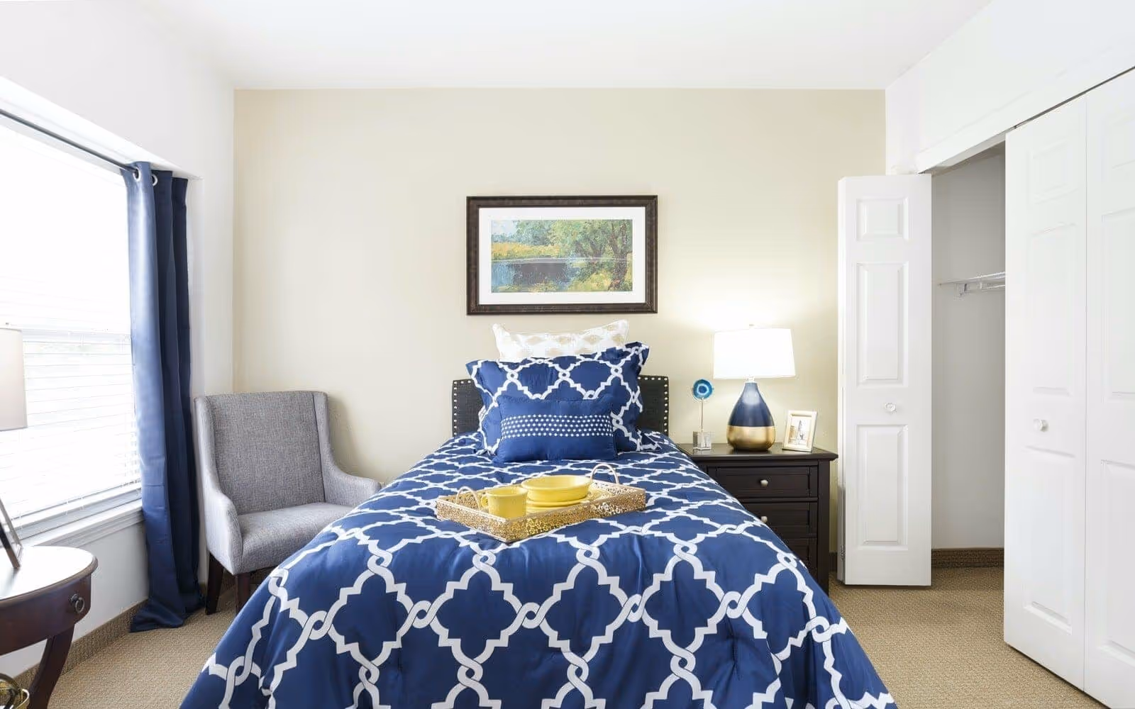 A bright bedroom with a single bed covered in a blue and white patterned comforter and matching pillows. A tray with yellow dishes is placed on the bed. To the right of the bed is a dark wooden nightstand with a blue and gold lamp, a small framed photo, and a decorative item. A framed landscape painting hangs on the beige wall above the bed. To the left is a window with blue curtains and a gray upholstered chair. On the right side of the room is an open closet with white bi-fold doors.