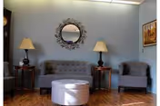 A cozy sitting area with a tufted sofa, two armchairs, side tables with lamps, a round wall mirror, and an ottoman on hardwood flooring.