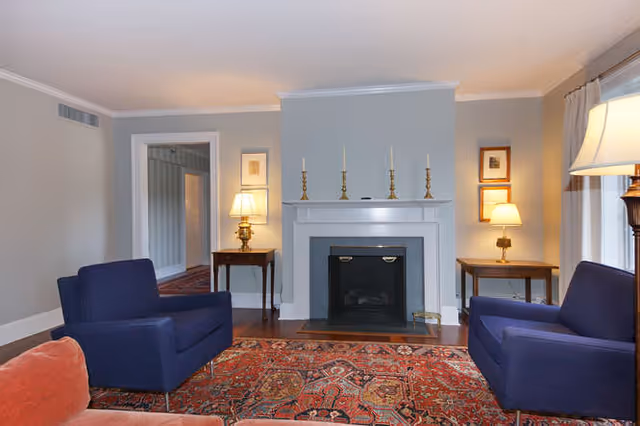 A cozy living room with two blue armchairs and a red patterned rug facing a white fireplace with four brass candlesticks on the mantel. There are two wooden side tables with lamps on either side of the fireplace, and a large window with curtains on the right side of the room.