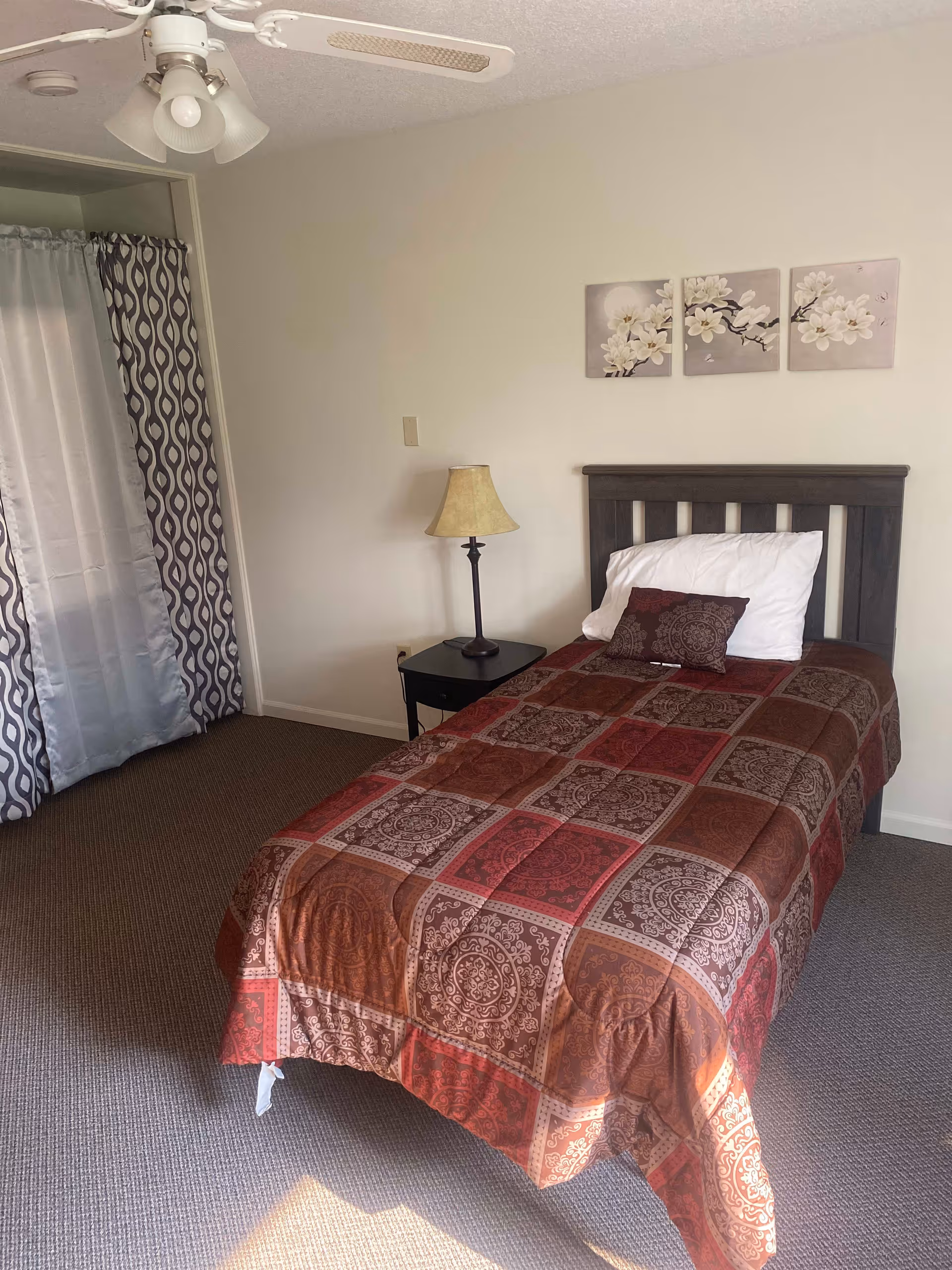 A small bedroom with a single bed covered in a patterned red and brown quilt. There is a white pillow and a matching decorative pillow on the bed. Next to the bed is a black nightstand with a beige lamp. Above the bed are three wall art panels featuring white flowers on a light background. A ceiling fan with lights is visible on the ceiling. The room has carpeted flooring and a window with patterned curtains.