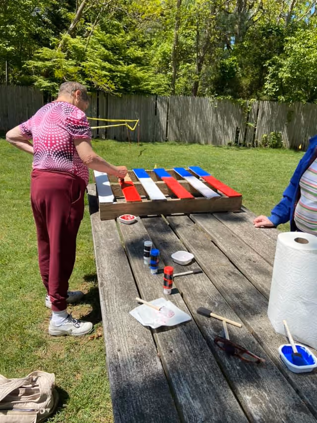 Two people painting a wooden pallet with red, white, and blue stripes on a picnic table in a grassy backyard.