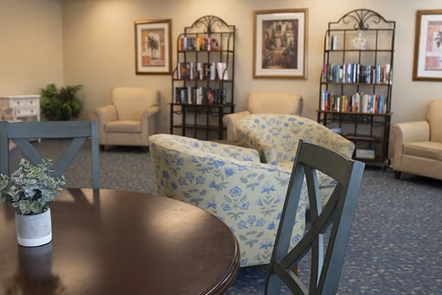 A cozy common area with a round wooden table and chairs in the foreground, two floral upholstered armchairs, beige armchairs, two black metal bookshelves filled with books, framed artwork on the walls, and a small plant on a white cabinet in the background.