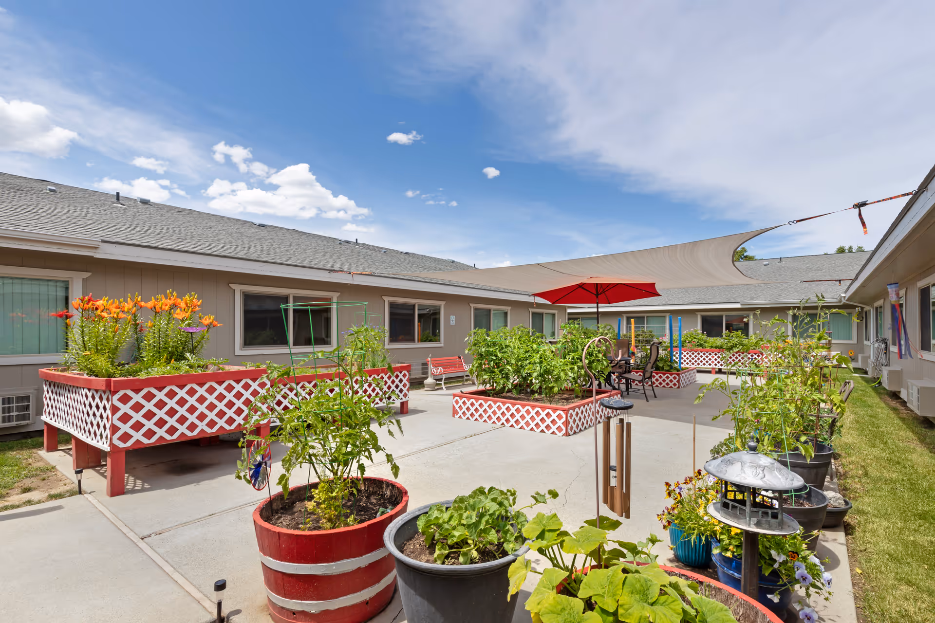 Sunny courtyard with raised planter boxes, potted plants, patio seating and a red umbrella surrounded by a single-story building.