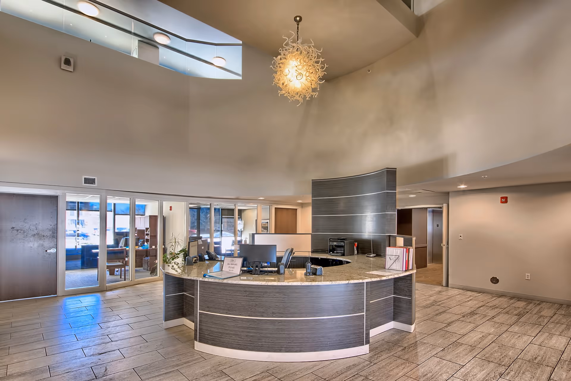 A modern reception area with a curved desk featuring computer monitors, office supplies, and a sign. The space has high ceilings, a decorative hanging light fixture, tiled floors, and glass doors leading to other rooms.