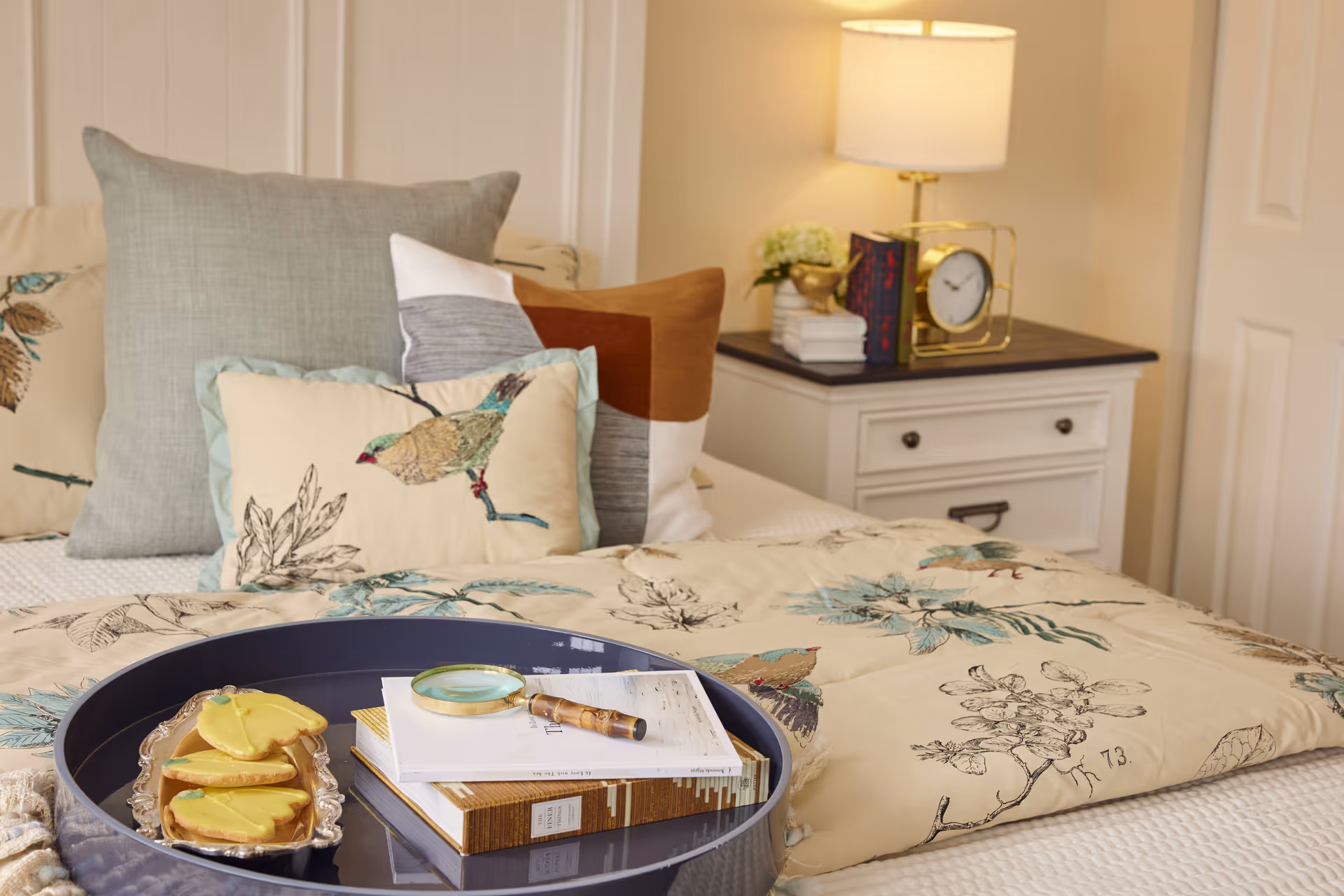 A cozy bedroom scene with a bed adorned with multiple decorative pillows, including one with a bird design. On the bed is a dark blue tray holding a magnifying glass, two books, and a small plate with yellow butterfly-shaped cookies. A white nightstand with drawers is beside the bed, topped with a lamp, a clock, some books, and a small plant.