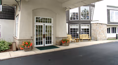 Entrance to a senior living facility with double glass doors framed by stone and beige walls. There are two wooden benches and a small table on the right side of the entrance. Flower pots with red flowers are placed on either side of the door. The building has large windows and a paved driveway in front.