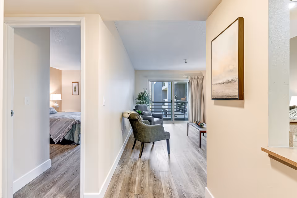 View down a hallway in a retirement center apartment showing a bedroom on the left with a bed and lamp, and a living room straight ahead with two gray armchairs, a coffee table, a potted plant, and a sliding glass door leading to a balcony. The walls are light-colored and the floor has wood-style flooring.