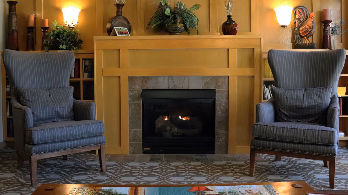 A cozy living room area with two striped armchairs facing a lit fireplace. The fireplace has a wooden mantel decorated with plants, vases, candles, and a rooster figurine. The walls are painted a warm yellow, and there are built-in shelves with books and decorative items on either side of the fireplace.